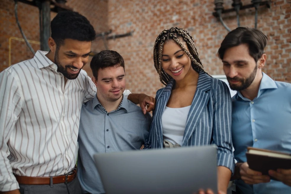 Disabled man with down syndrome with 4 colleagues at work all looking at laptop and smiling